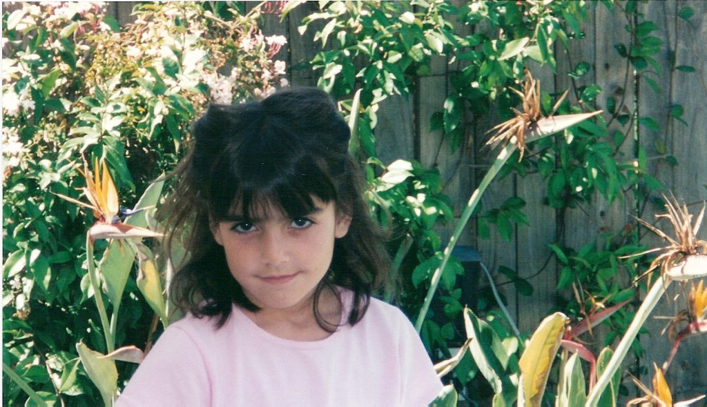 the artist at maybe 6 years old, in a pink shirt and hair done up, surrounded by bird of paradise flowers 